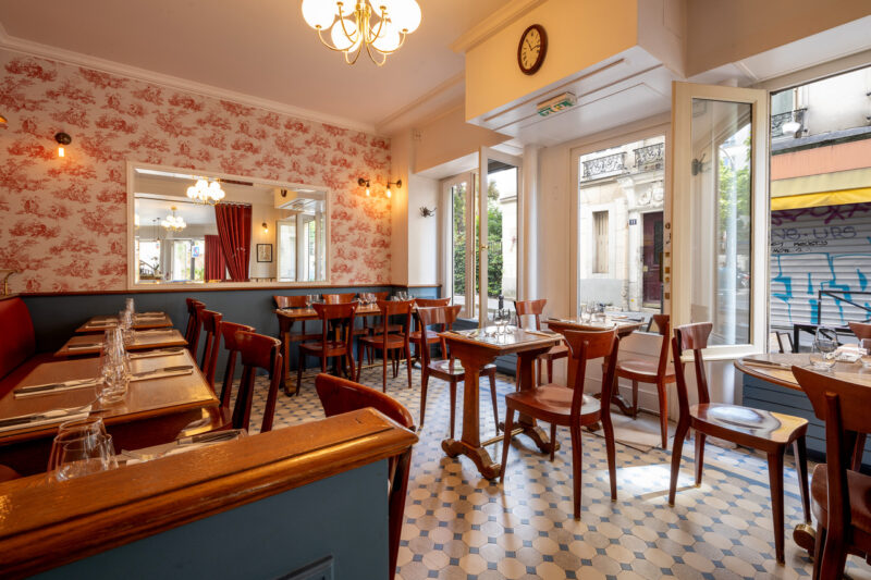 Cozy restaurant interior with floral wallpaper, wooden tables, and chairs, featuring a clock and large windows.