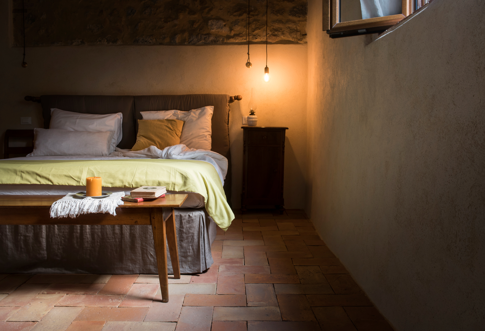 Cozy bedroom with a bed covered in a yellow blanket, wooden bench, and soft lighting from a hanging bulb.