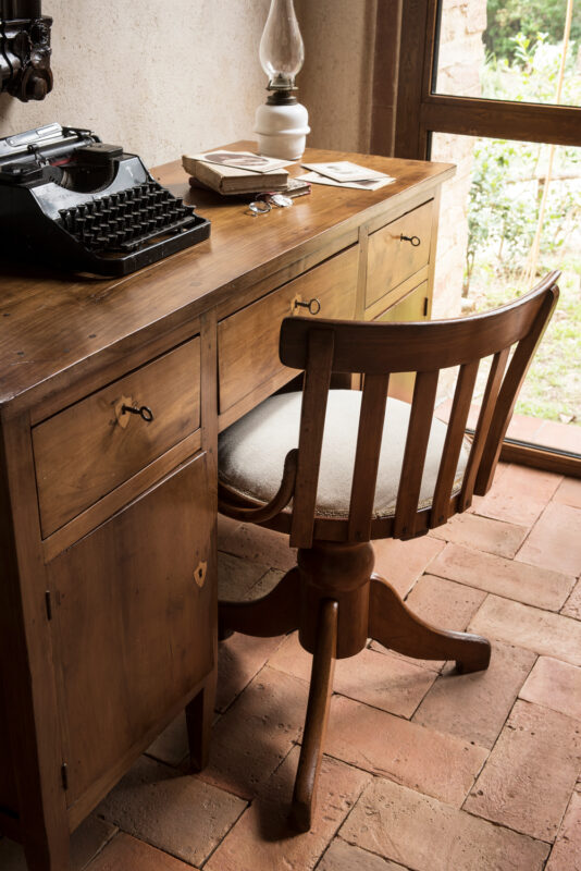 Wooden desk with a typewriter, books, and a lamp, beside a window overlooking greenery.