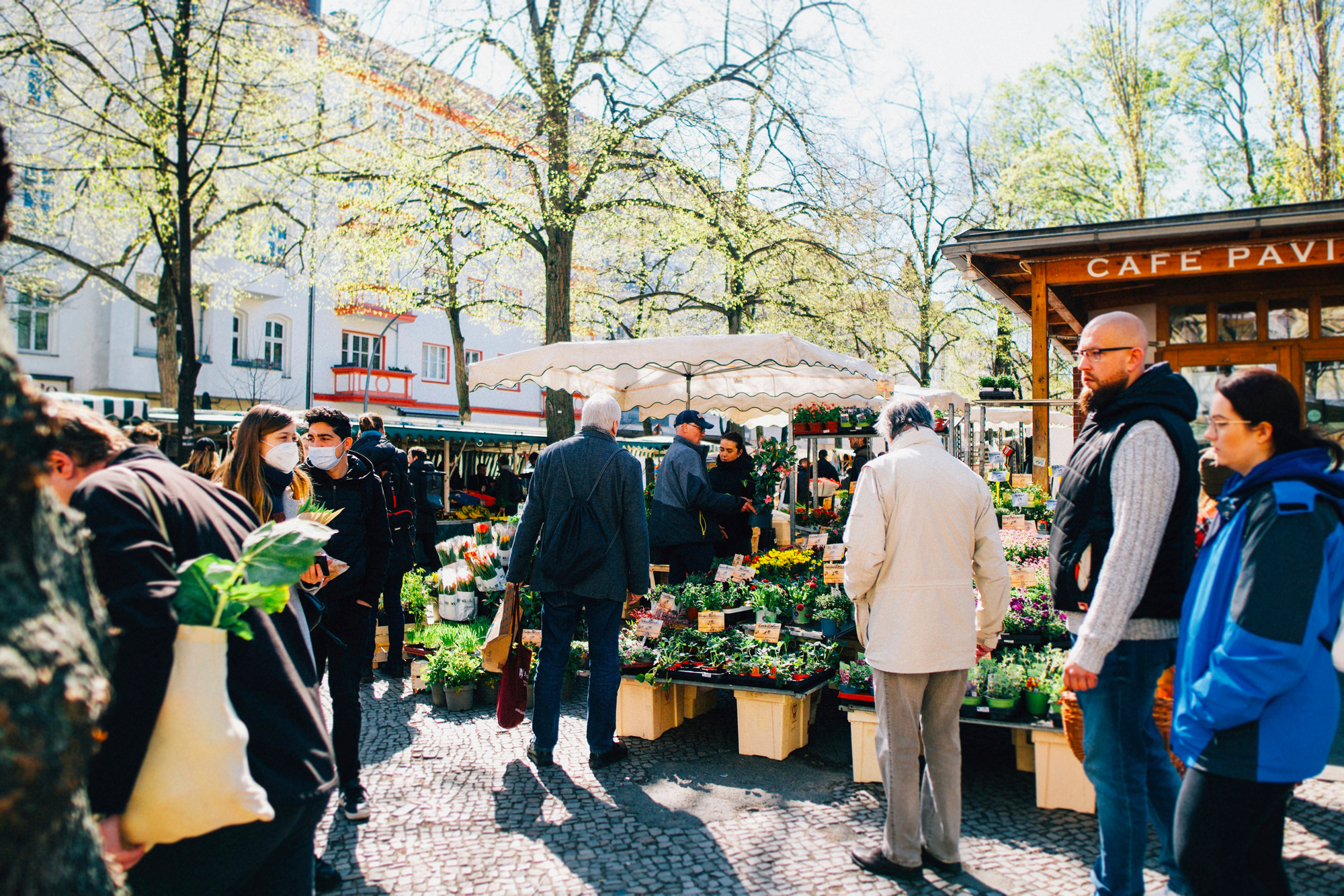 Busy outdoor flower market with people browsing stalls, surrounded by trees and a café in the background.