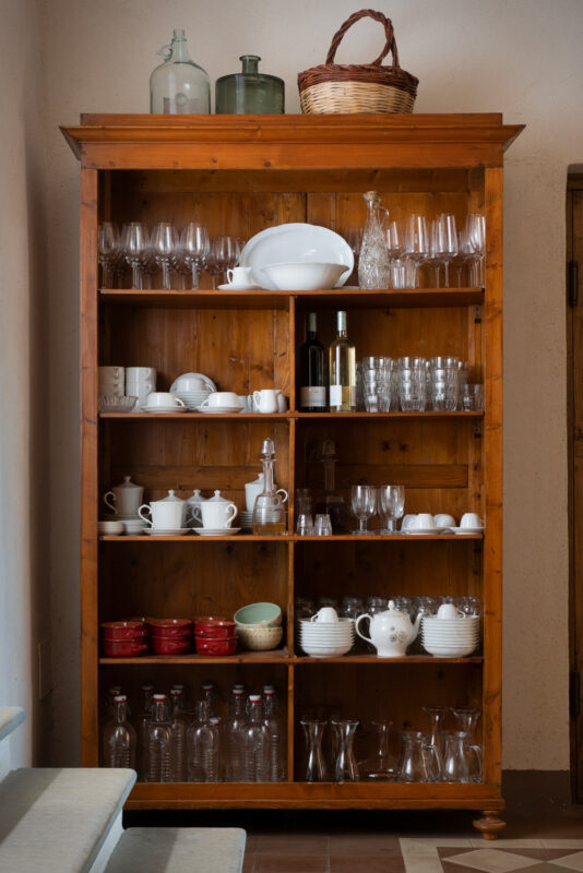 Wooden shelving unit filled with various glassware, dishes, and a basket, set against a light wall.