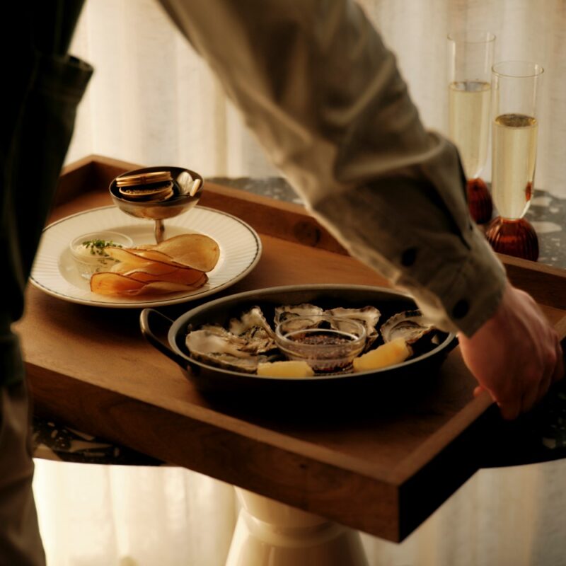A waiter serves a tray with oysters, lemon, and a plate of chips, alongside two glasses of champagne.