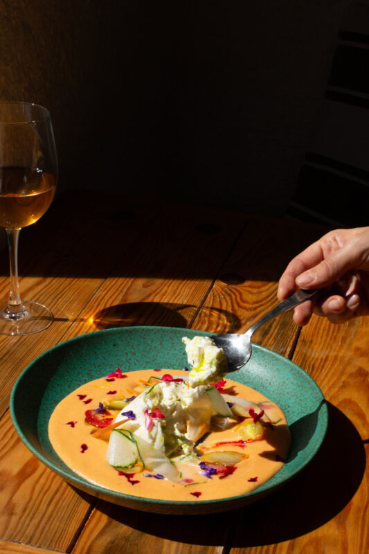 A hand holding a spoon scoops creamy gazpacho topped with edible flowers, beside a glass of white wine on a wooden table.