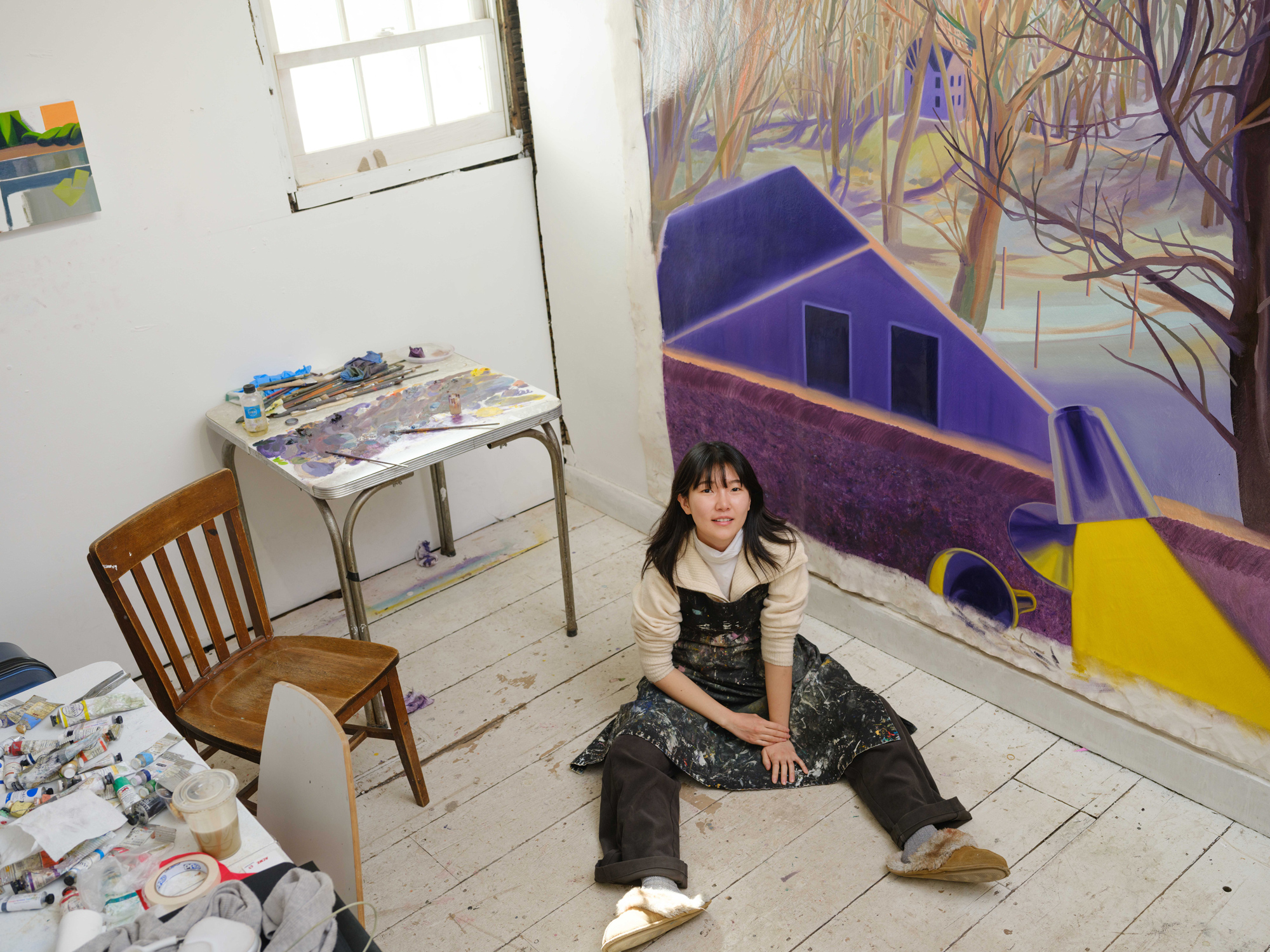 Heejo Kim sits on the floor of her studio, surrounded by art supplies, with a colorful mural in progress on the wall.