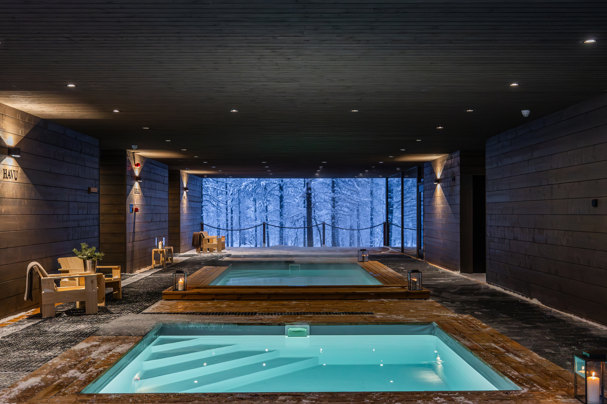 Indoor spa area with two hot tubs, surrounded by wooden walls and a snowy forest view through large windows.