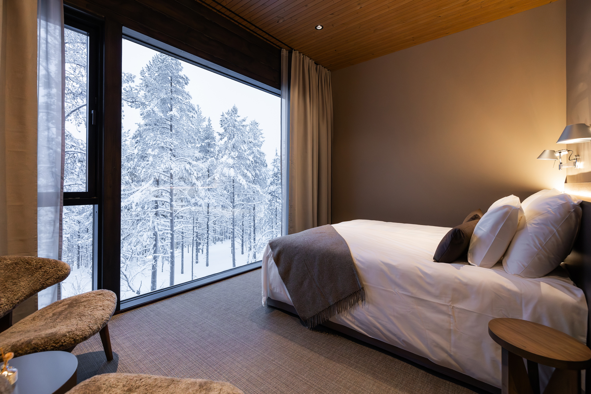Cozy hotel room with a bed, two chairs, and a large window showcasing a snowy forest landscape.