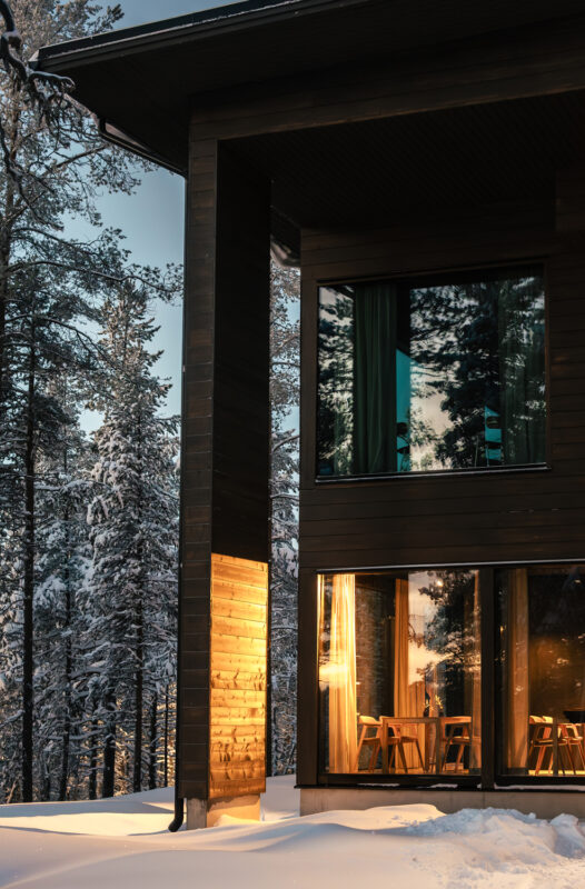 Modern wooden hotel exterior with large windows reflecting snowy trees, illuminated by warm light at dusk.