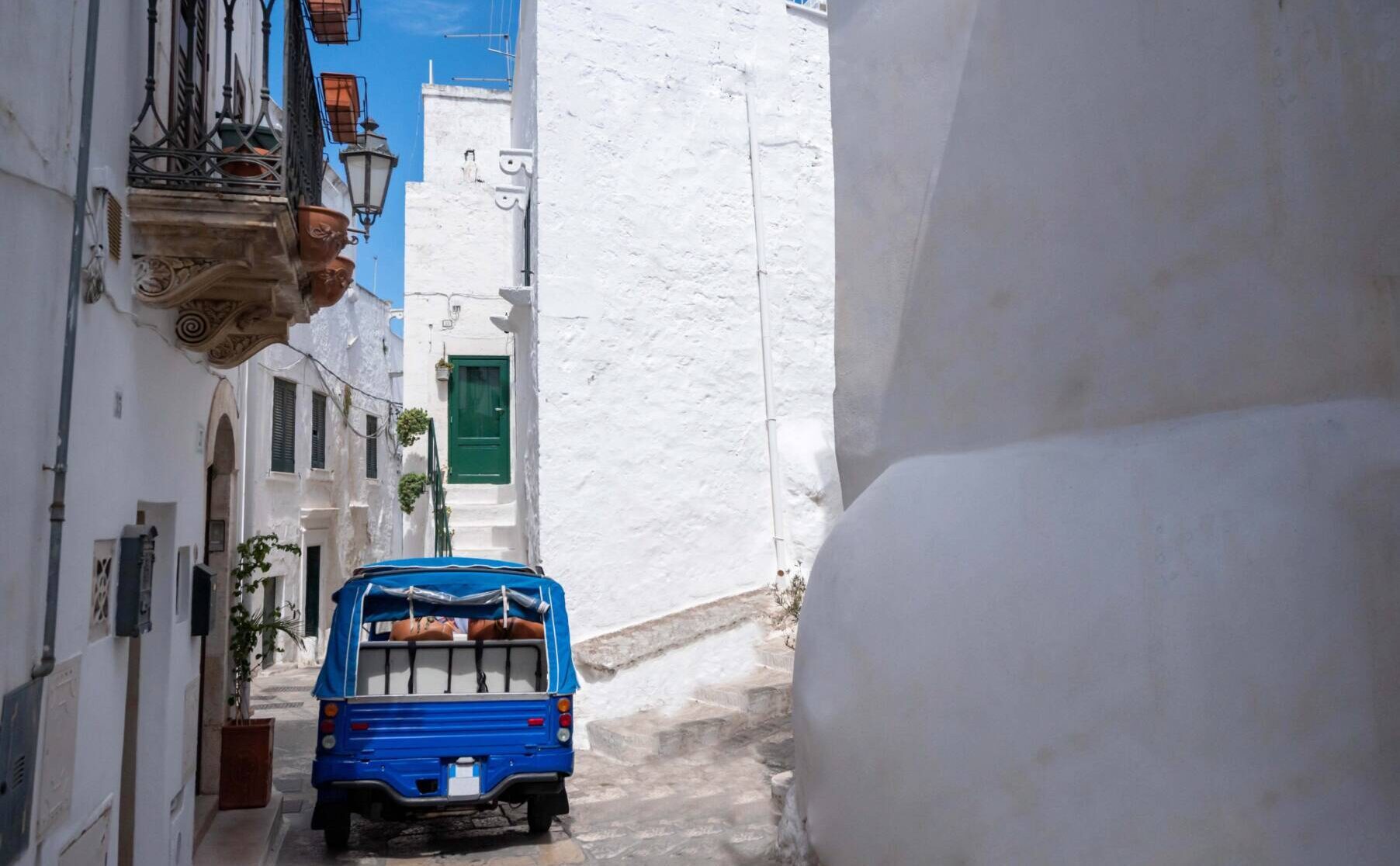 Narrow street in Ostuni with a blue vehicle parked beside whitewashed buildings and a green door.