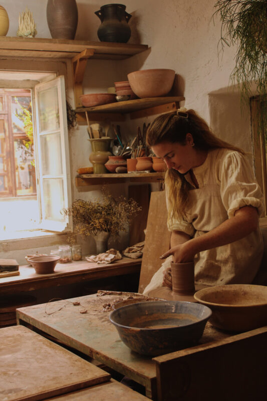 A woman in a light-colored dress shapes clay on a potter's wheel in a sunlit pottery studio, surrounded by various pottery...