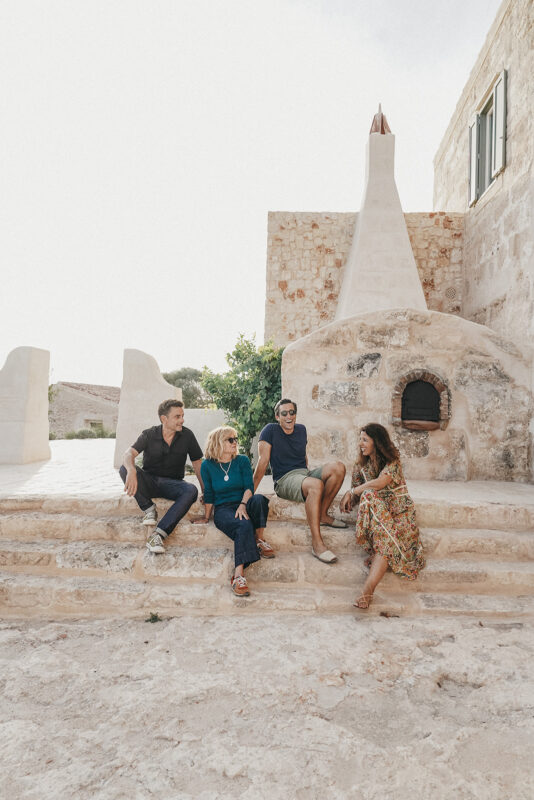 Four people sitting on stone steps outside a rustic building in Son Blanc, Menorca, enjoying a sunny day.