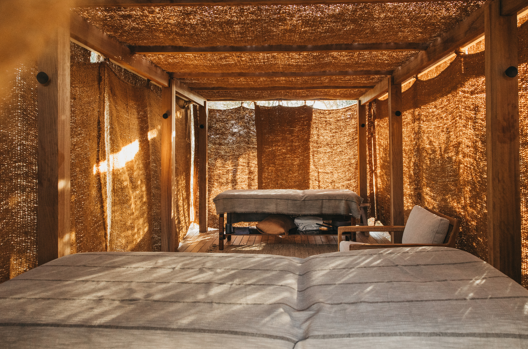 Spa treatment area with wooden beds and a chair, surrounded by woven fabric walls in warm tones.