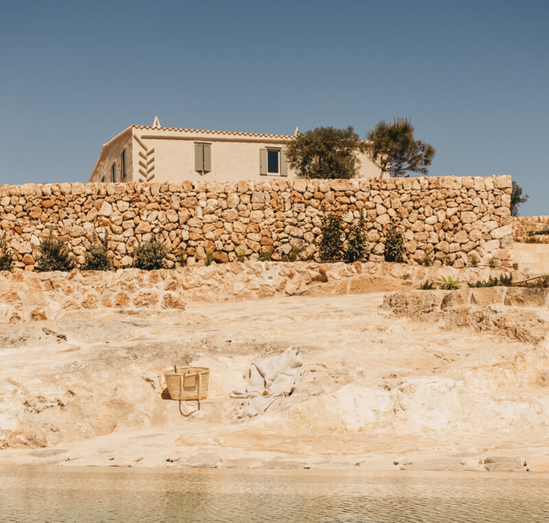 Stone walls and a rustic building overlook a tranquil pool in a sunlit landscape at Son Blanc, Menorca.
