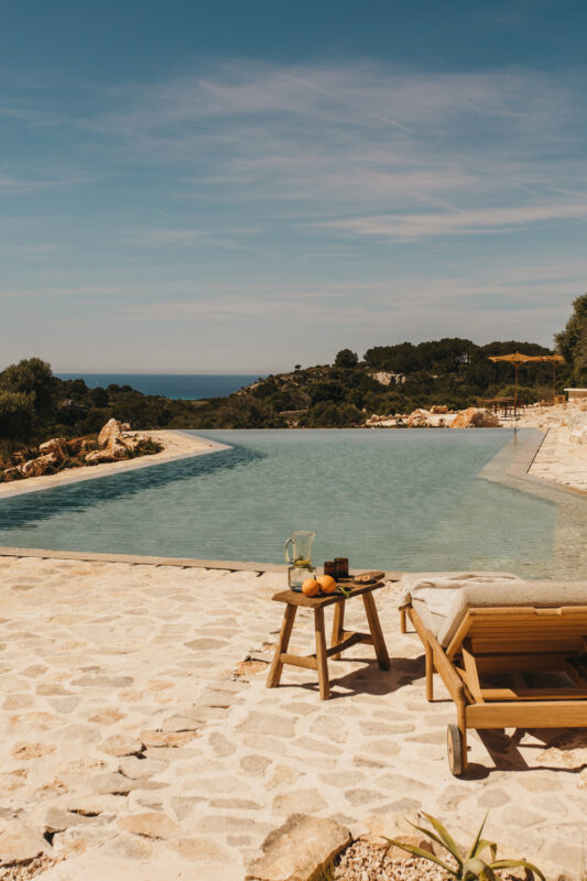 Infinity pool with a stone deck, a wooden chair, and a small table with fruit, overlooking a scenic landscape and sea.