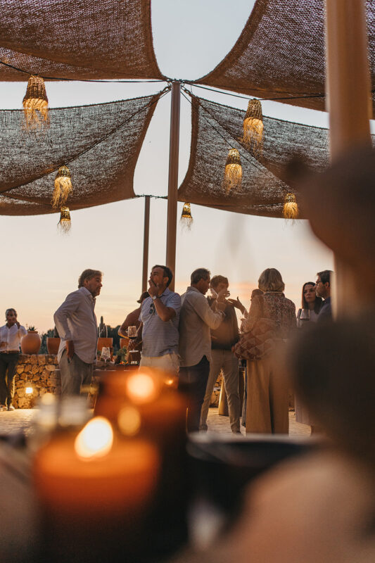 Evening gathering under woven shade with people socializing, soft lighting from candles in the foreground.