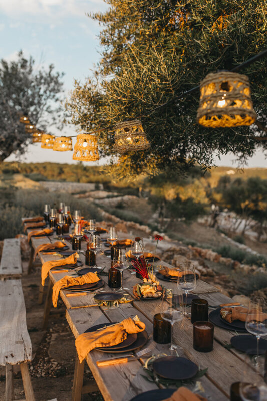 Elegant outdoor dining setup with wooden table, black plates, and amber napkins, adorned with hanging lanterns and greenery.