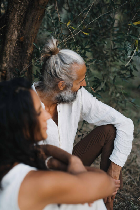 A man with long gray hair and a beard sits beside a woman with dark hair, both in a natural setting among trees.