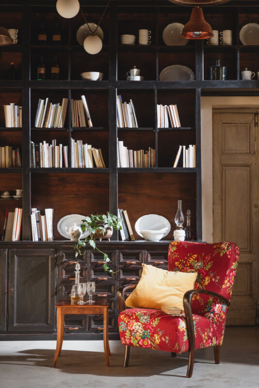 Cozy living room with a red floral armchair, wooden shelves filled with books and dishes, and a vintage wooden table.