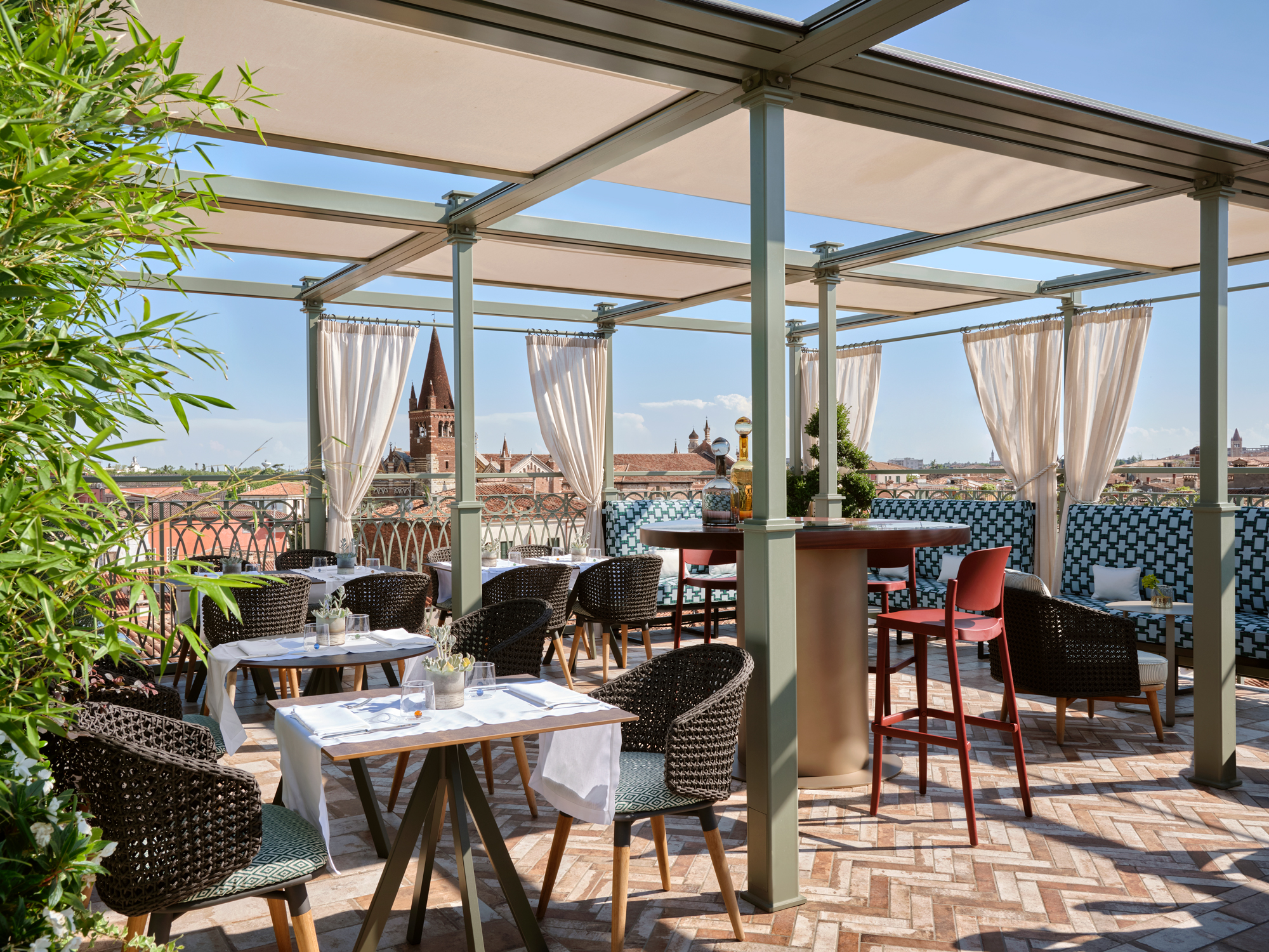 Rooftop terrace in Verona with wicker chairs, tables, and draped curtains, overlooking city rooftops and a church spire.