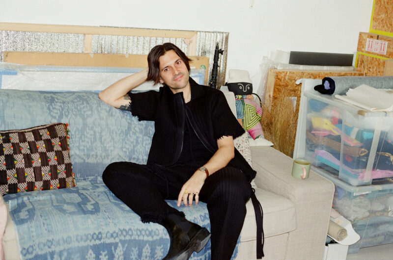 Man sitting on a couch in a studio, wearing a black outfit, with colorful art and storage boxes in the background.