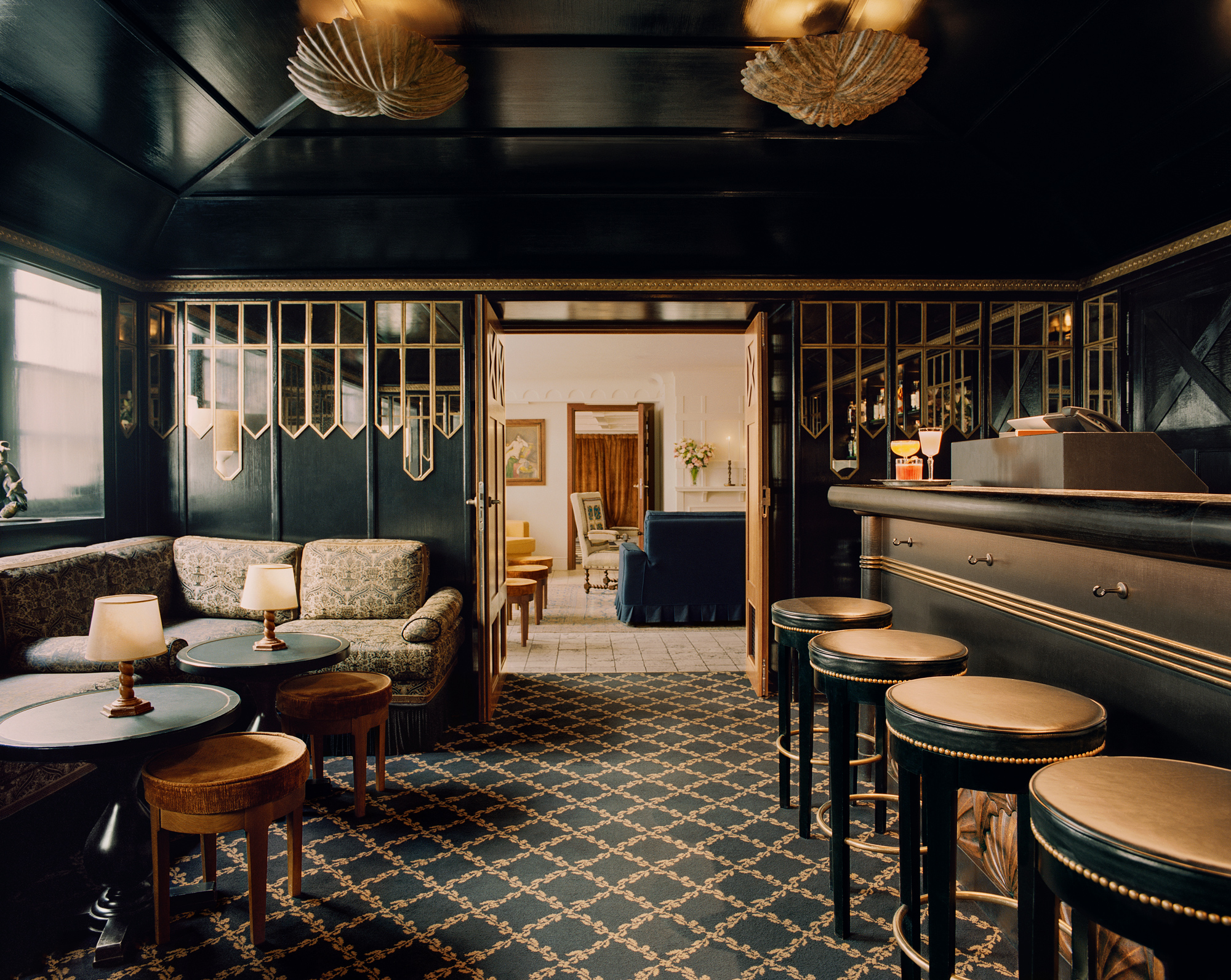 Elegant interior of a bar area with patterned carpet, dark walls, and stylish seating, leading to a bright room beyond.