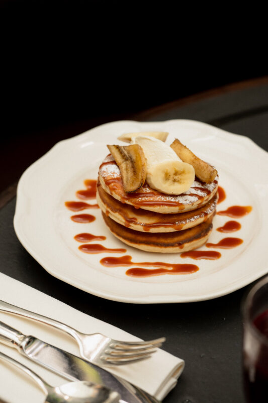 Stack of pancakes topped with banana slices and drizzled with sauce, served on a white plate with cutlery beside it.
