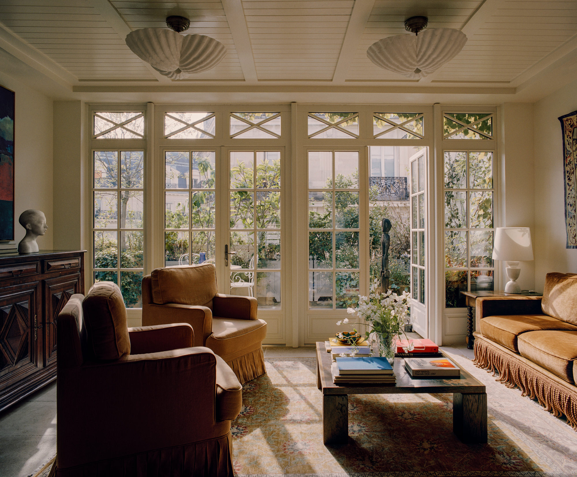 Bright living room with large windows, two armchairs, a sofa, and a coffee table adorned with books and flowers.