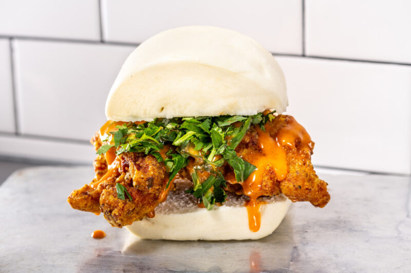 Fried chicken sandwich in a steamed bun topped with cilantro and sauce, set against a tiled background.