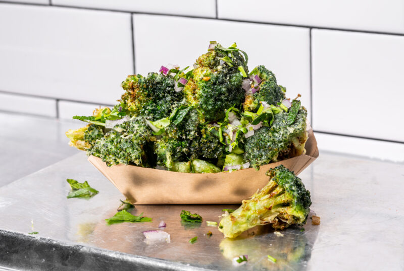 Tempura-battered broccoli topped with chopped herbs and onions in a brown paper tray on a stainless steel surface.