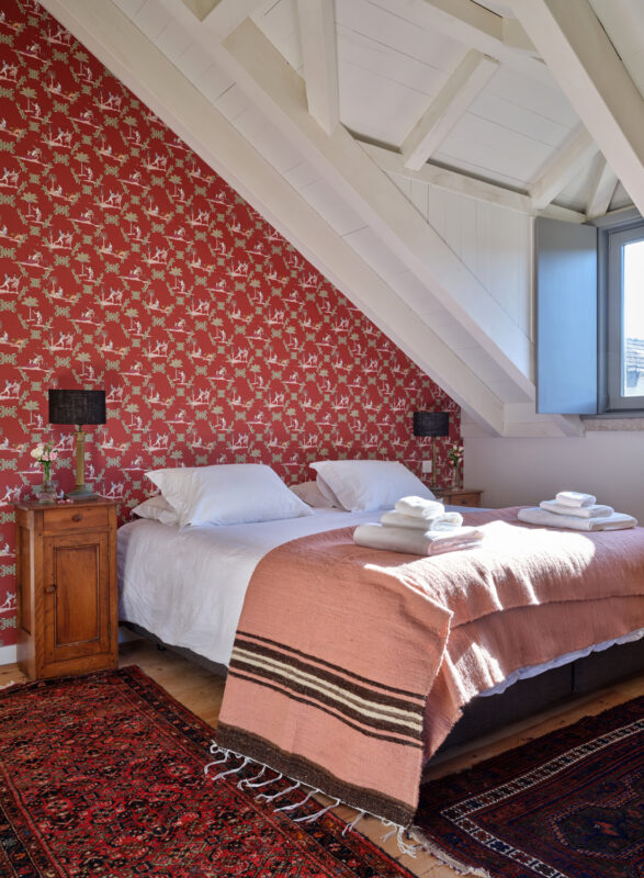 Cozy bedroom featuring a red patterned accent wall, two beds with white linens, and a pink blanket, illuminated by natural...