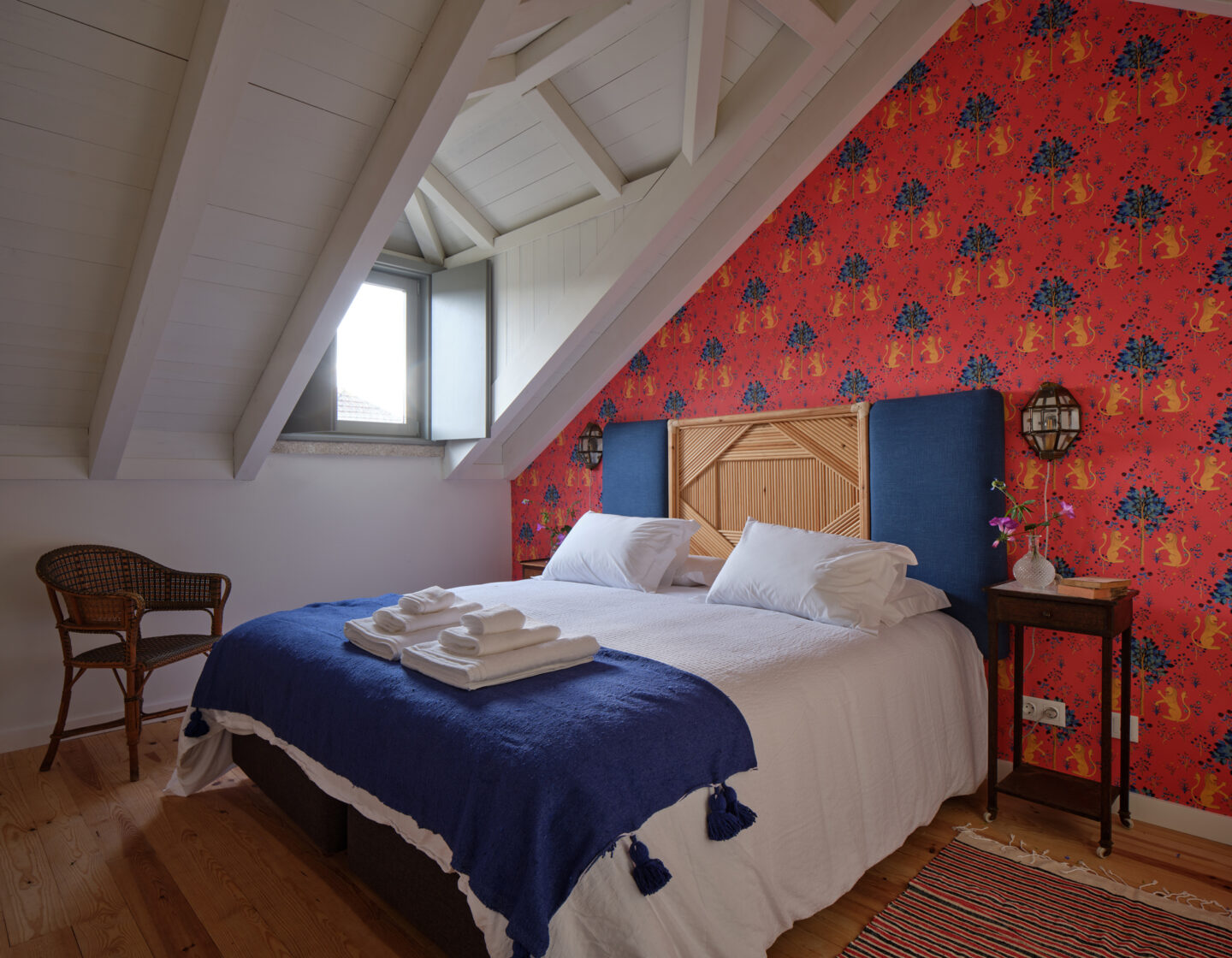 Cozy bedroom with a sloped ceiling, featuring a double bed with white linens, blue throw, and vibrant floral wallpaper.