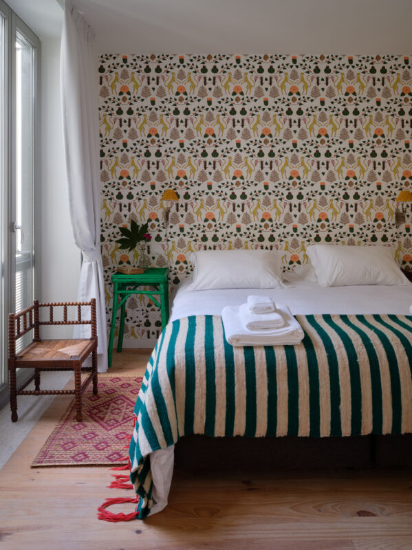 Cozy bedroom featuring a striped blanket, two beds, a patterned wallpaper, a small chair, and a green side table.