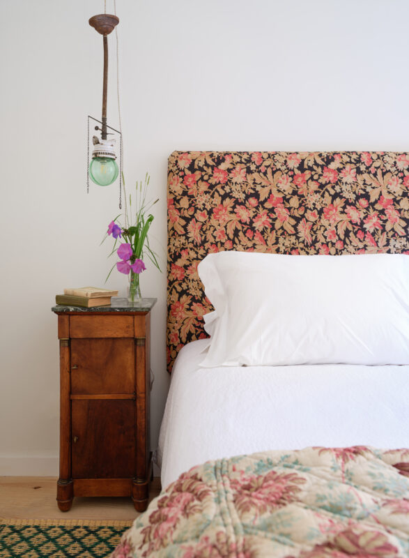 Cozy bedroom with a floral headboard, white bedding, a wooden nightstand, and a vase of flowers.