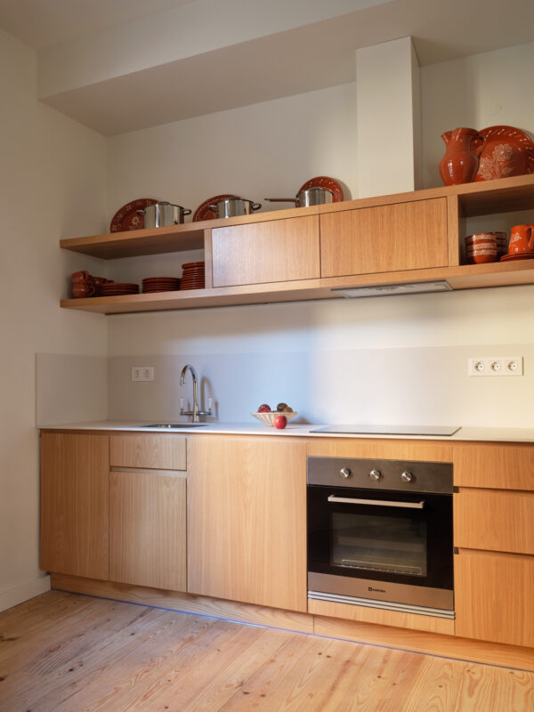 Modern kitchen with wooden cabinetry, stainless steel oven, and red ceramic dishware on shelves. Bright natural light from...