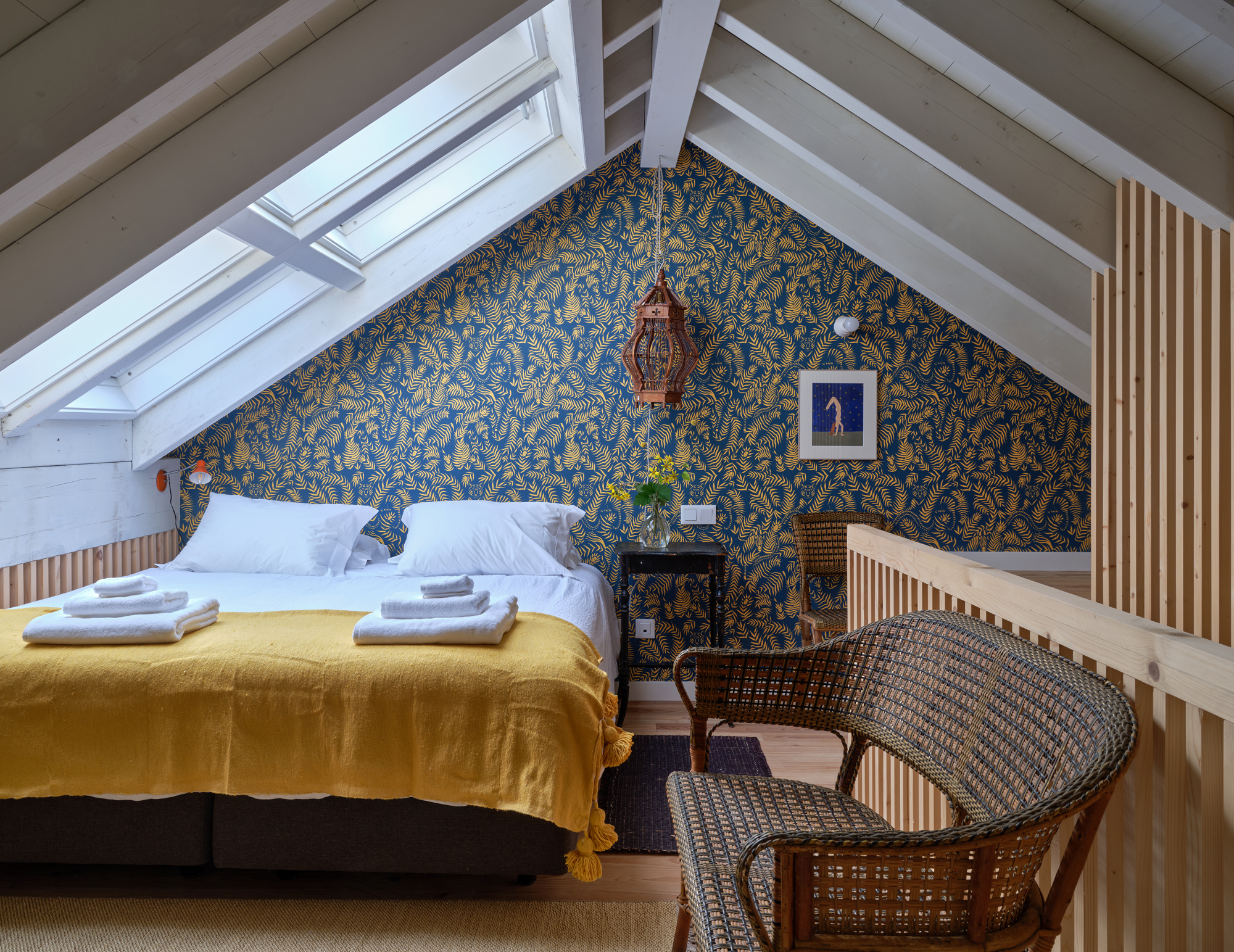 Cozy attic bedroom with a yellow blanket, two beds, patterned wallpaper, and a wicker chair.