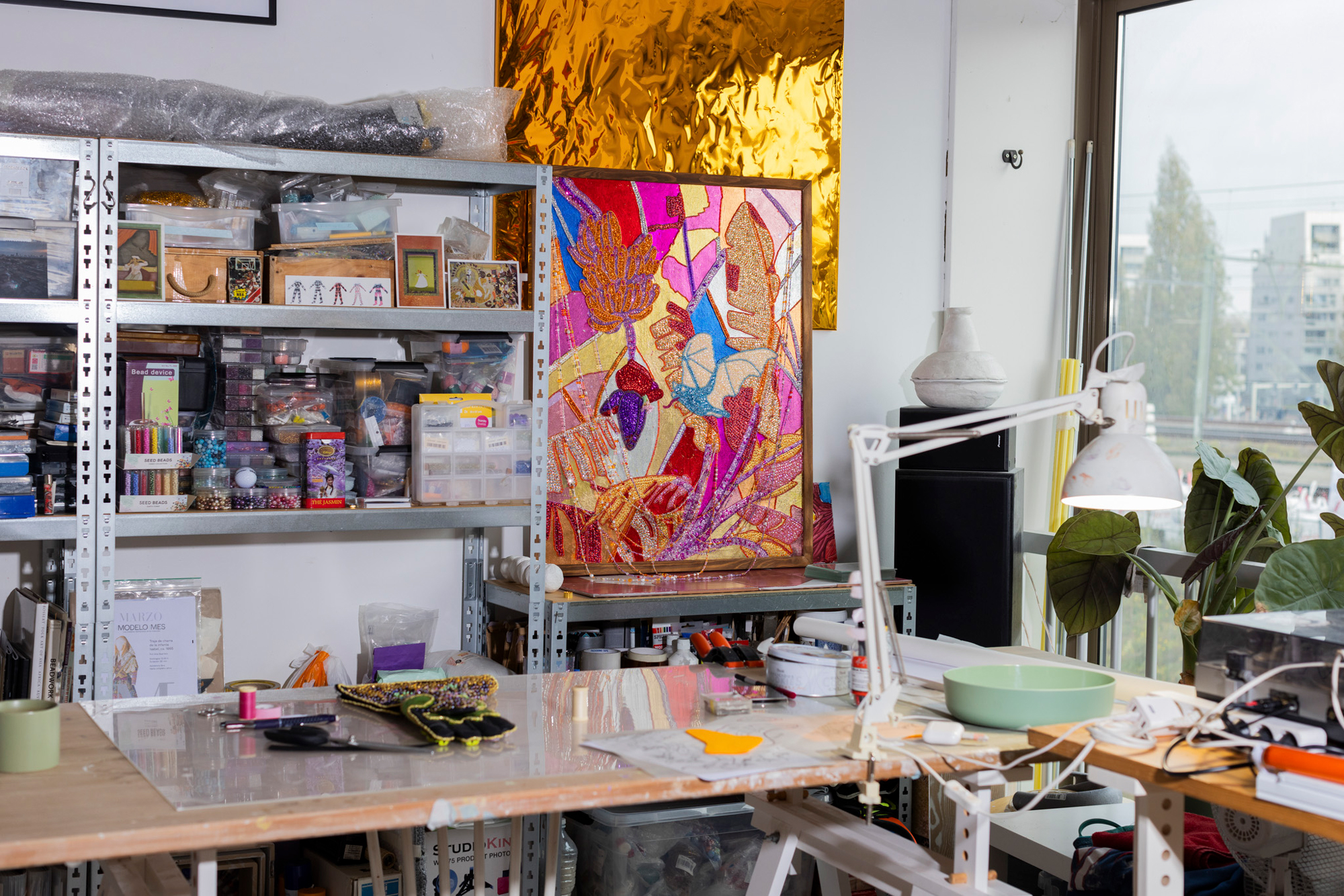 Art studio featuring a colorful artwork on display, cluttered shelves with various materials, and a work table.