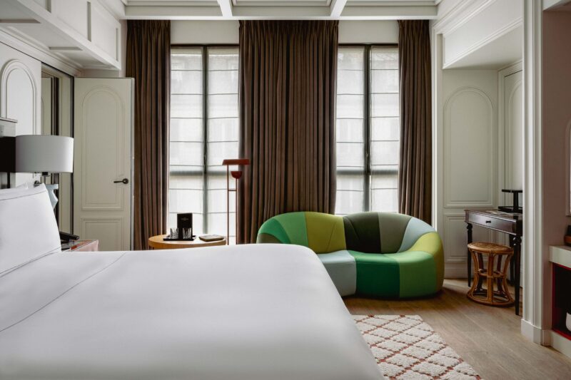Modern hotel room featuring a white bed, green and blue curved sofa, and large windows with curtains.