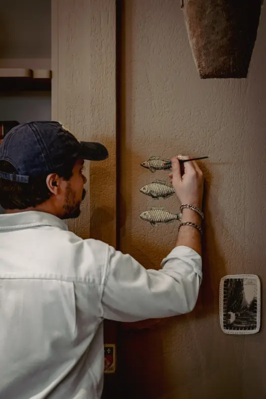 Person painting three fish on a textured wall, with a small framed picture and a hanging planter nearby.