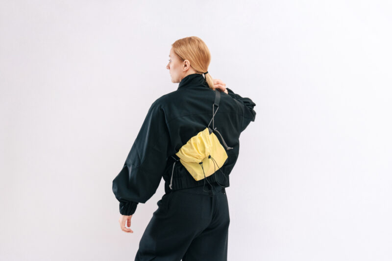Woman in a black jacket with a yellow pouch on the back, styled with a ponytail, against a white background.