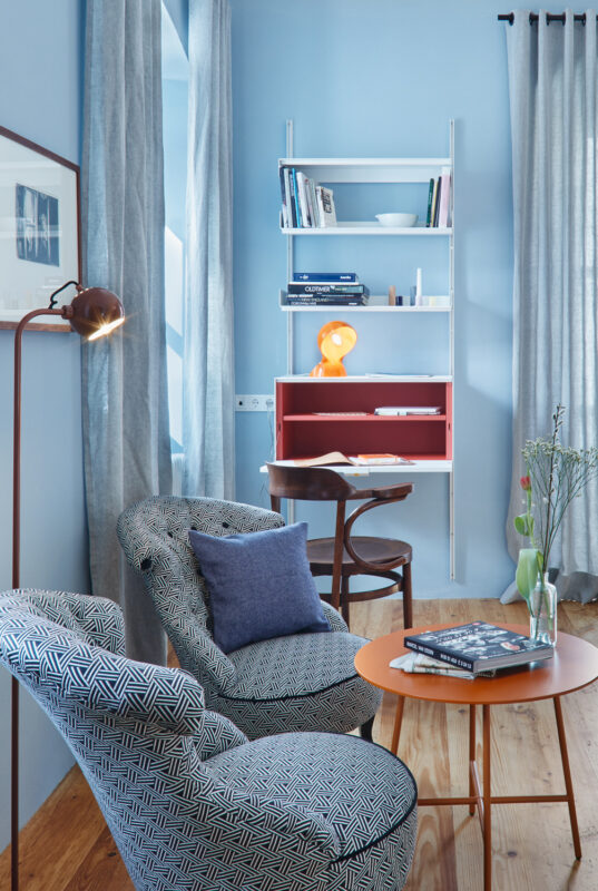 Cozy hotel room corner with patterned chairs, a small table, books, and a modern lamp against blue walls.