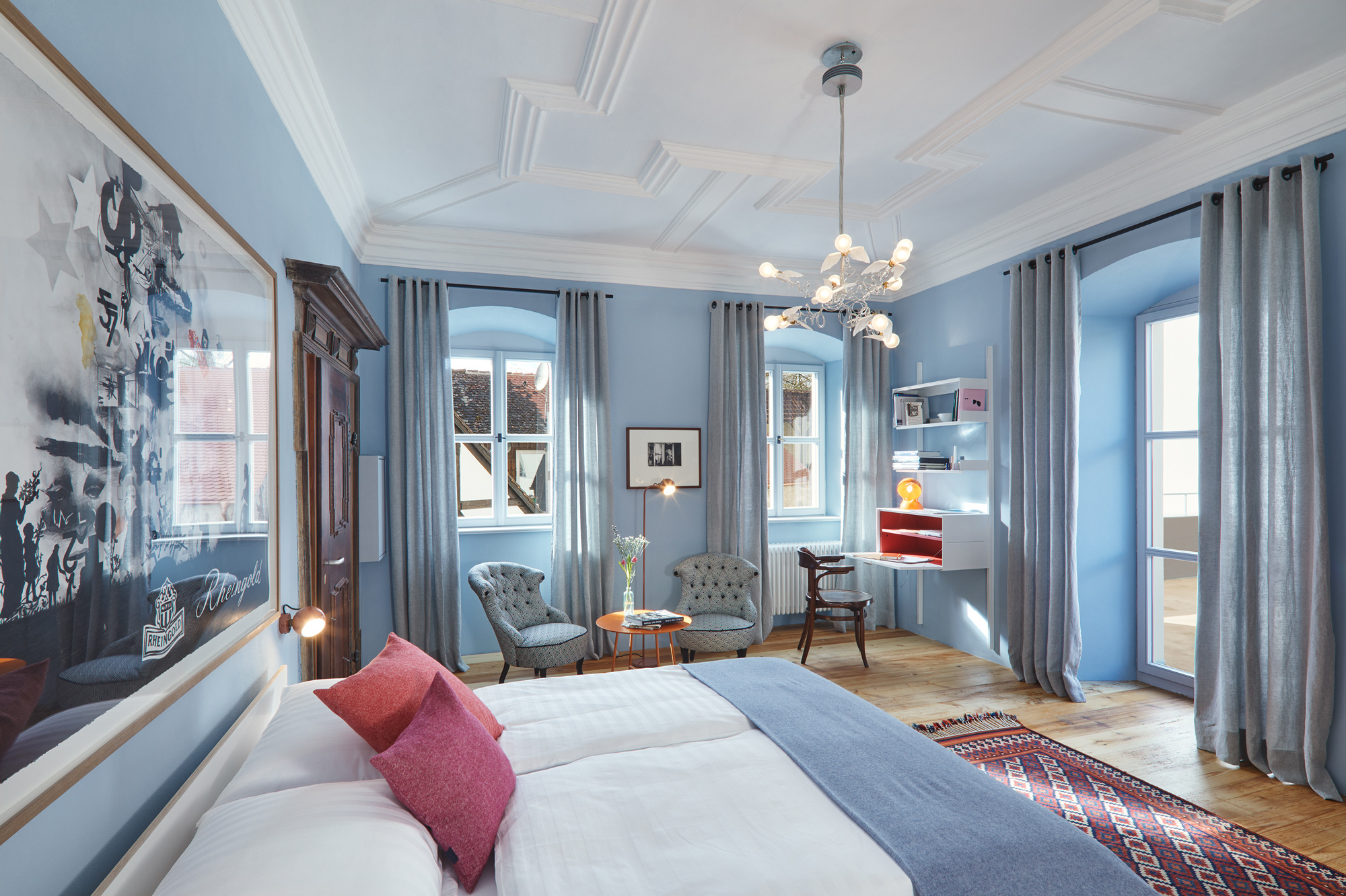 Bright hotel room with blue walls, a double bed, armchairs, a desk, and large windows, featuring modern decor and artwork.