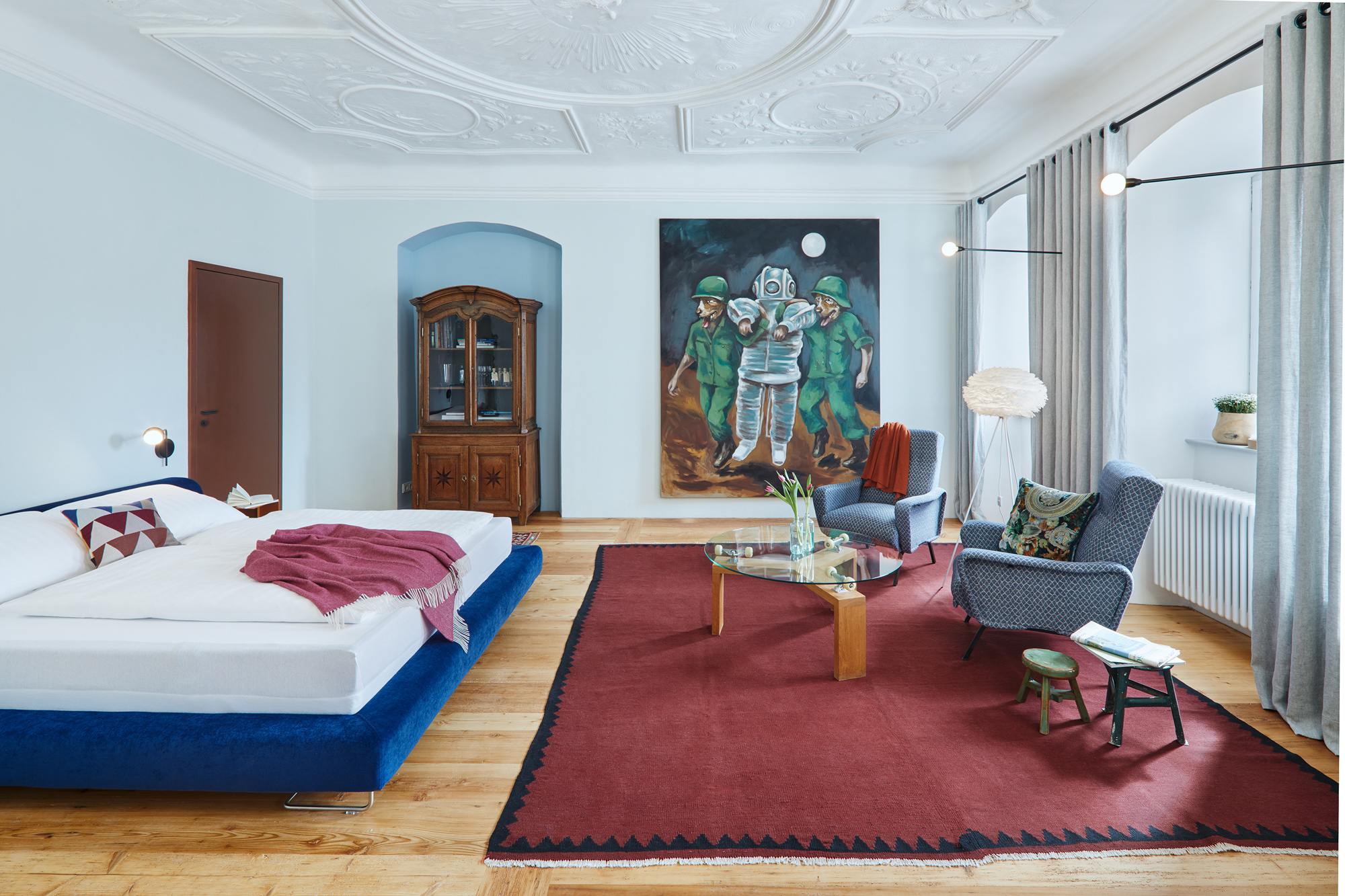 Stylish hotel room featuring a blue bed, wooden furniture, a large painting, and a red rug.