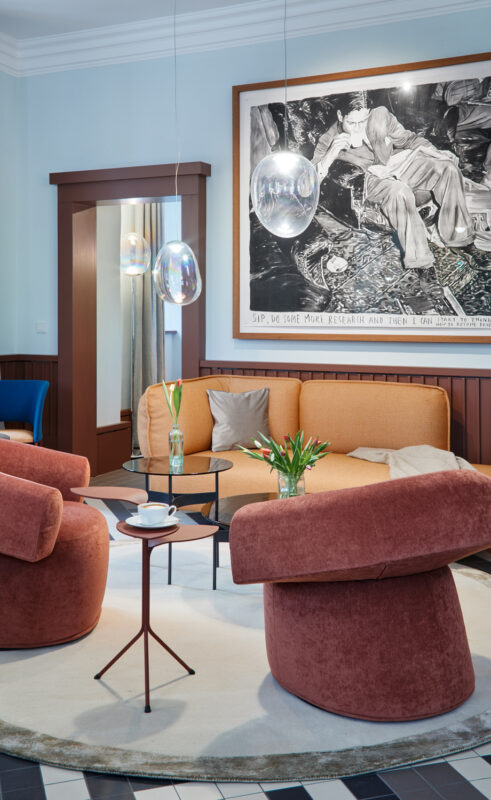 Cozy hotel lounge with orange and pink seating, a glass table, and modern pendant lights, featuring a large artwork on the...