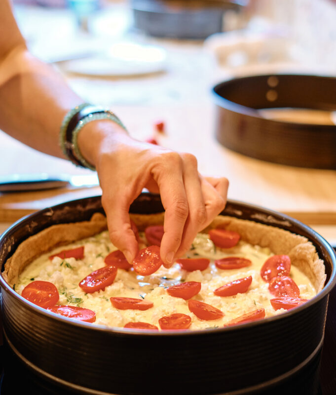Hand placing sliced tomatoes on a creamy dish in a black baking pan, with a kitchen setting in the background.