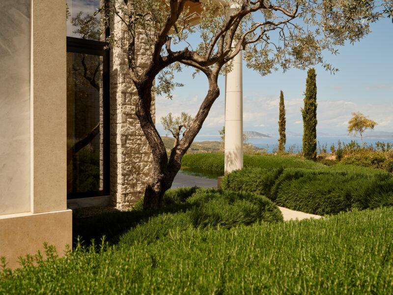 Lush greenery surrounds a stone structure, with a view of the sea and distant hills under a clear blue sky.