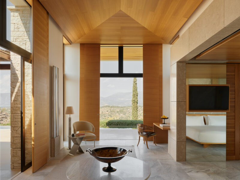 Spacious bedroom in Amanzoe, Greece, featuring wooden accents, large windows, and a view of the landscape.