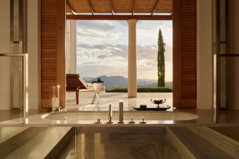 Luxurious pool pavilion at Amanzoe, featuring a sunlit interior with a view of mountains and a cypress tree.