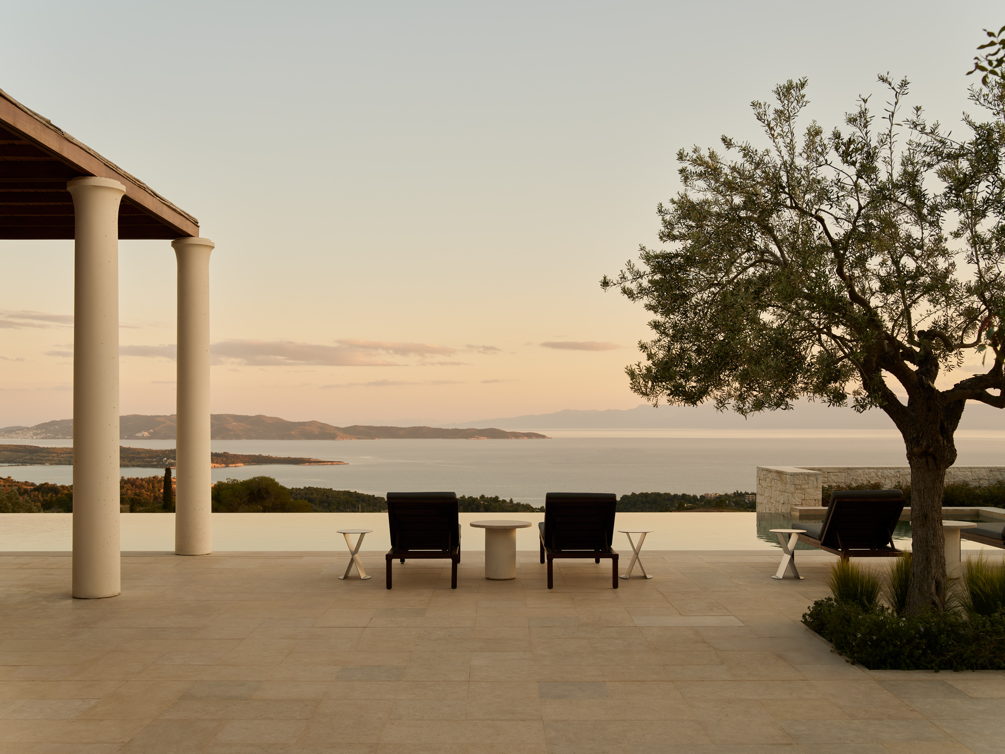 Outdoor area of Villa 2 at Amanzoe, featuring lounge chairs, a table, and a view of the sea at sunset.