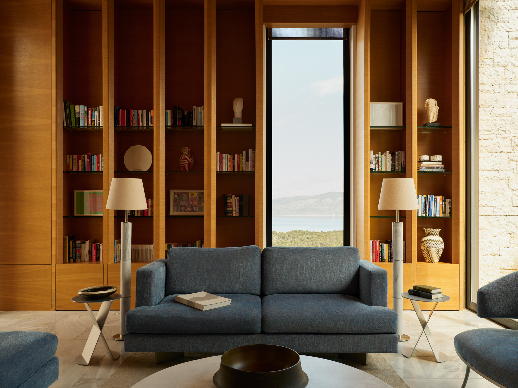 Modern living area in Villa 15 at Amanzoe, featuring a blue sofa, bookshelves, and a scenic view through a large window.