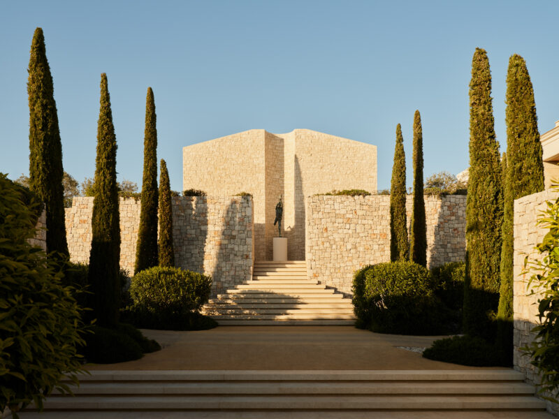 Stone steps lead to a modern, angular building surrounded by tall cypress trees and lush greenery at Amanzoe Resort, Greece.