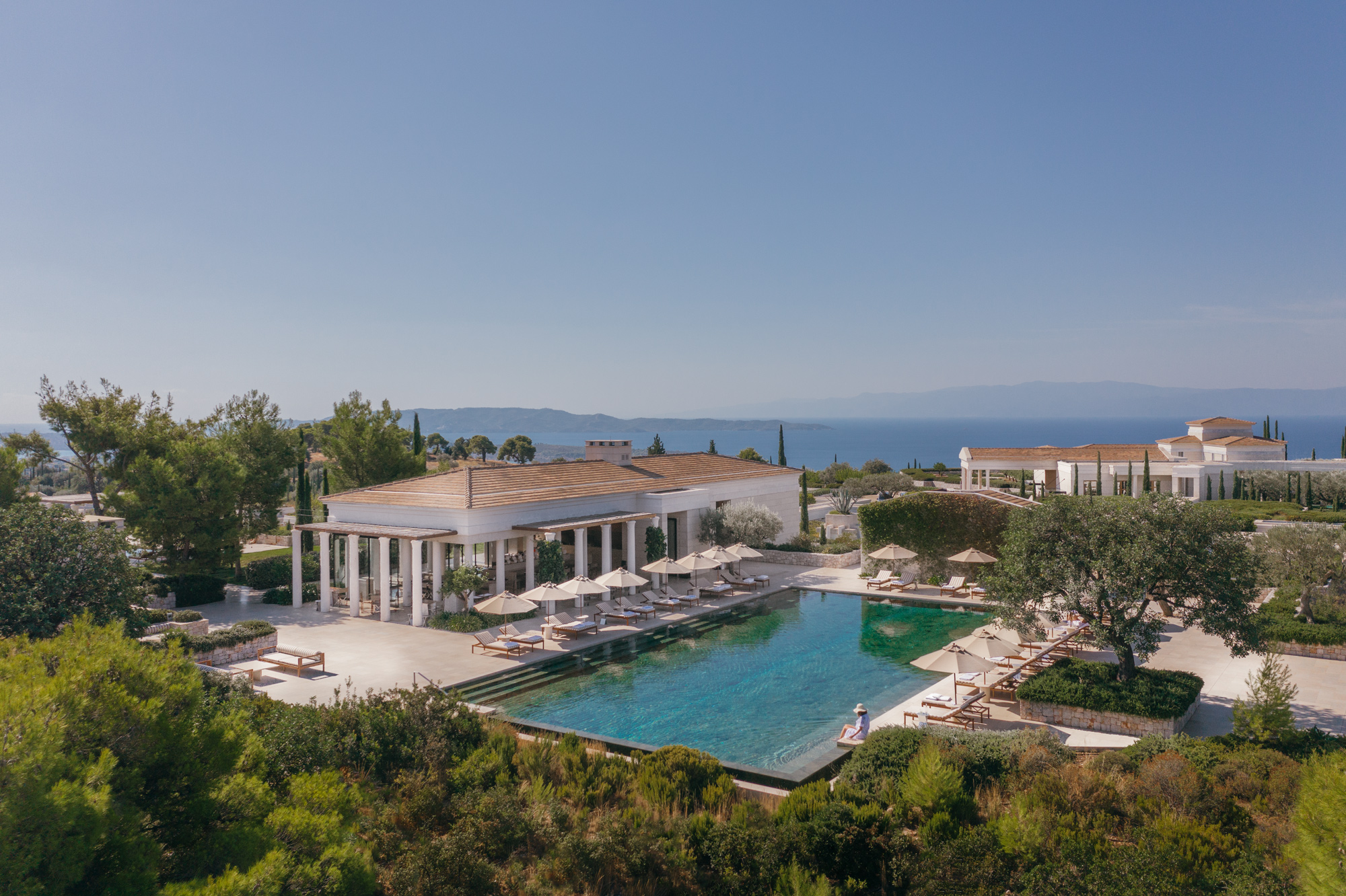 Aerial view of Amanzoe Resort in Greece, featuring a main swimming pool surrounded by sun loungers and lush greenery.